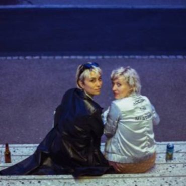 two blonde women seated on a staircase are looking behind